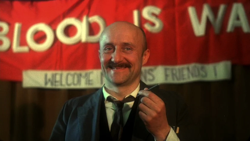 The War of the Worlds: Next Century (1981) movie still: medium — A man stands before a red banner with white text in a dimly lit hall. The man holds…