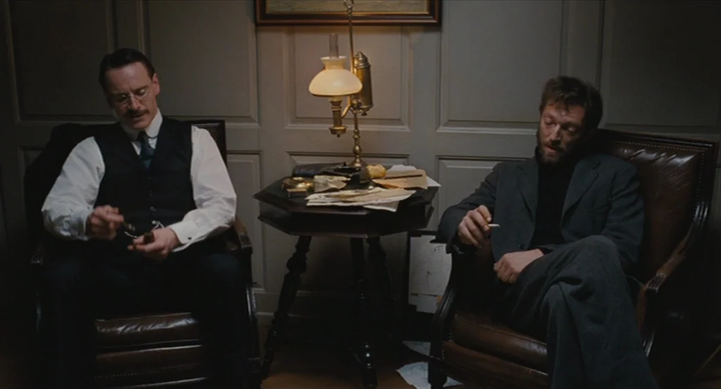 A Dangerous Method movie still: medium — A dark, wood-paneled room features two men seated in leather armchairs around a table. A small oil lamp…