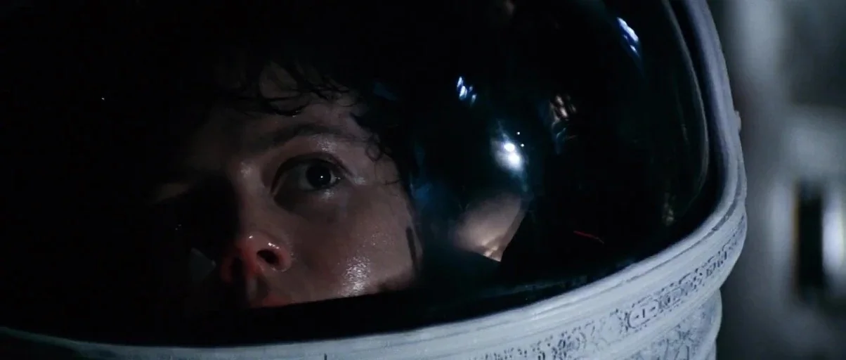 Alien (1979) movie still: closeup — A close up of a face peering from inside a white helmet visor. The dark interior shows a woman looking intently…