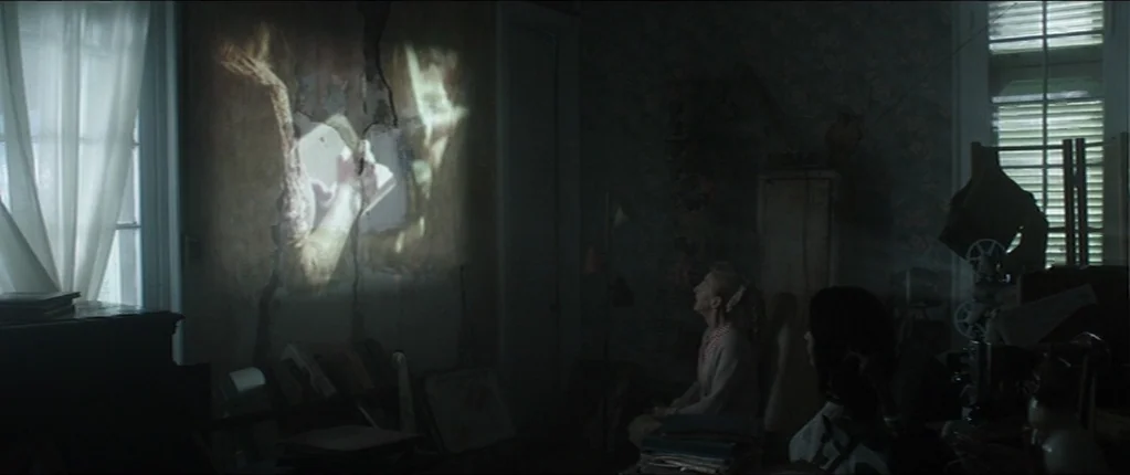 Abattoir (2016) movie still: wide — A dimly lit room contains stacks of books and a home movie projector. Two women sit facing a wall where a film…