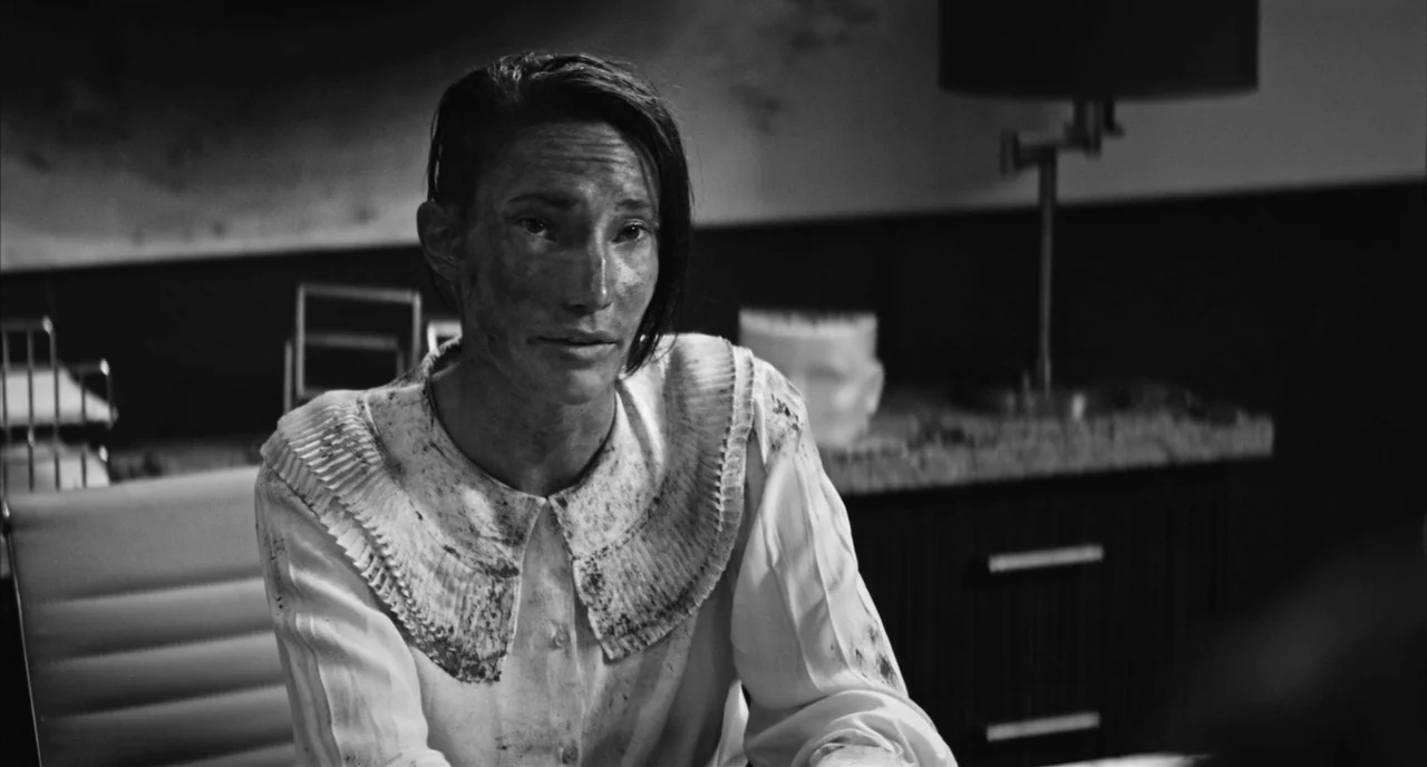 Nightmare Cinema movie still: medium — A dimly lit office features dark furniture and a desk with scattered items. A woman sits in the foreground…