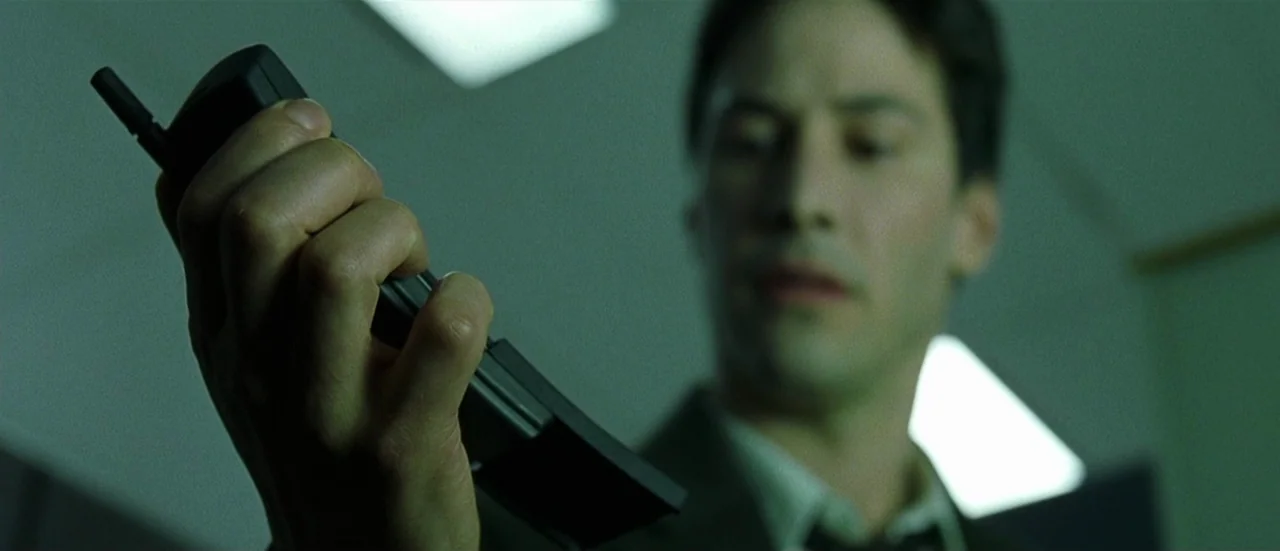 The Matrix (1999) movie still: closeup — A man in a suit looks down at a handheld device in his palm. The environment is lit with greenish…