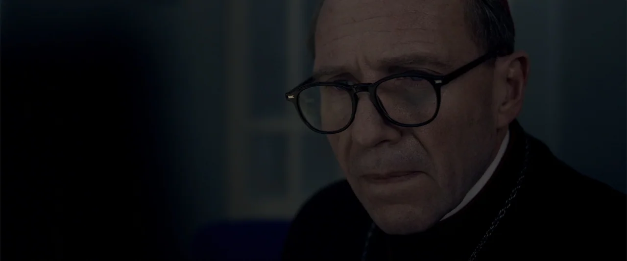 Conclave (2024) movie still: closeup — A dark office setting with muted blue tones and minimal architectural detail. An elderly man in clerical…