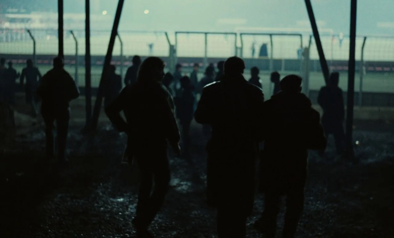 Graduate First (1978) movie still: wide — A dark muddy area outside a stadium fence at night. Several figures in heavy coats walk away from the…