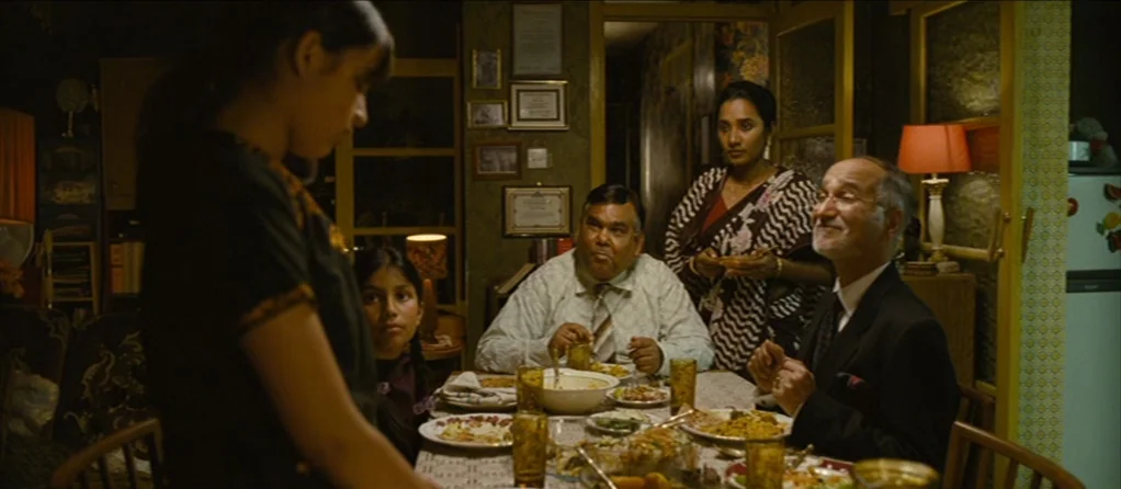 Brick Lane (2007) movie still: medium — A dimly lit dining area features multiple family members gathered around a table with plates. Framed…