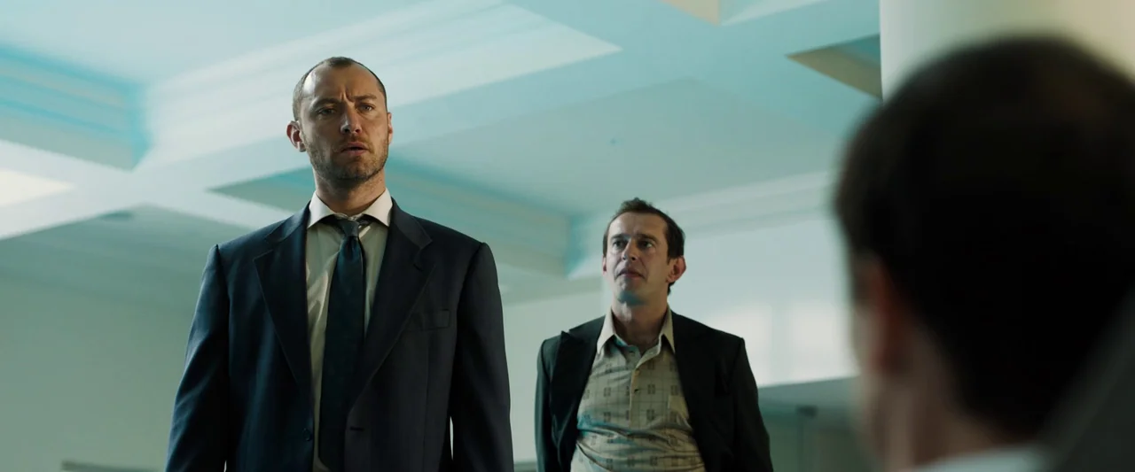 Black Sea (2014) movie still: medium — A modern office interior with a white coffered ceiling and diffuse lighting. Two men in business attire stand…