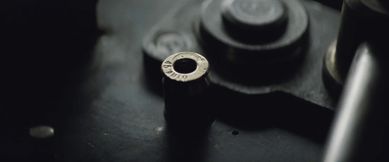Colt 45 (2014) movie still: closeup — A close-up view shows a .45 ACP shell casing sitting on a dark machine. The textured metallic surface of the…