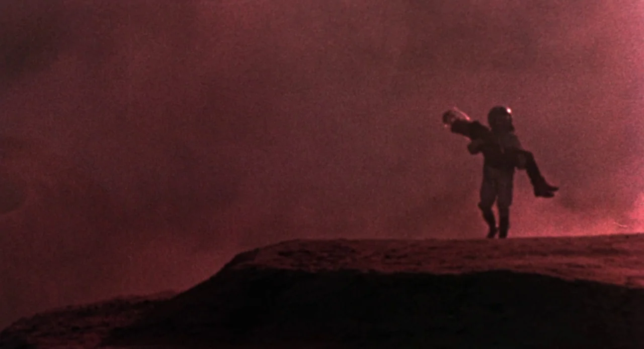 Queen of Blood (1966) movie still: wide — A desolate, red-lit terrain with a soft rise in the foreground and haze. A person in a white space suit…
