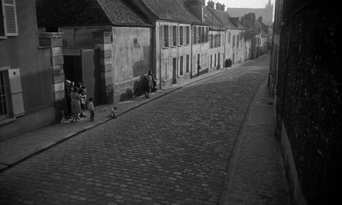Léon Morin, Priest (1961) movie still: wide — A narrow, sloping cobblestone street in a European town is lined with aged buildings. Several people…