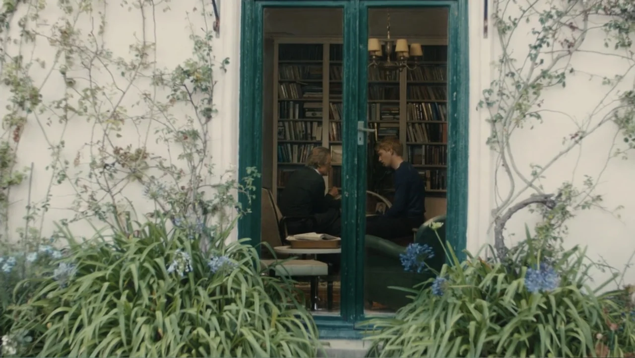 About Time (2013) movie still: medium — An interior home office seen through an exterior glass door framed by garden plants. Two men sit in…
