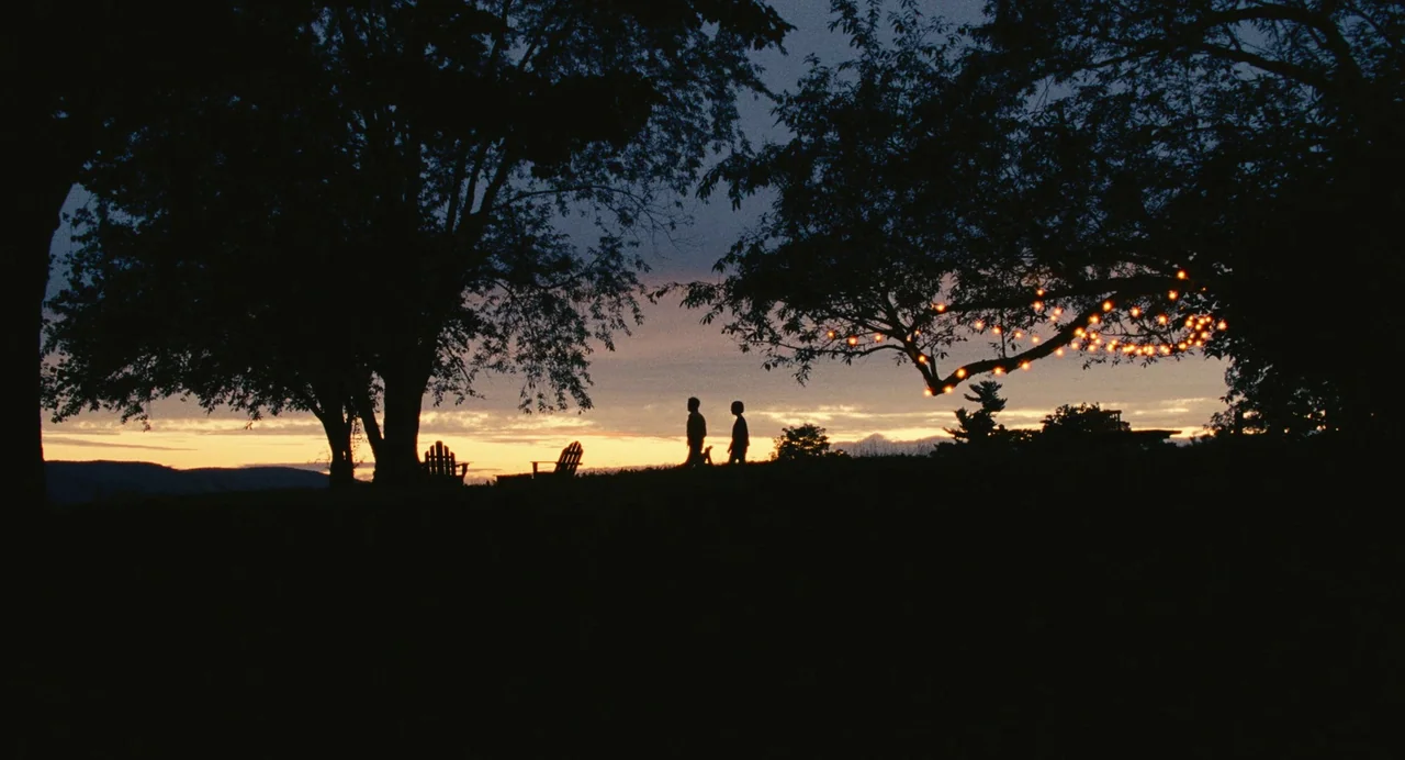 Past Lives (2023) movie still: wide — A grassy hilltop features large trees under a twilight sky with a colorful horizon. Two small figures walk…