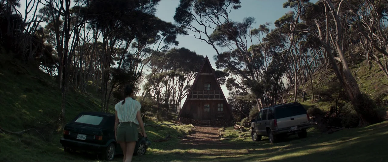 Evil Dead Rise (2023) movie still: wide — A secluded A-frame cabin sits in a clearing surrounded by tall, dense coniferous trees. A woman walks…