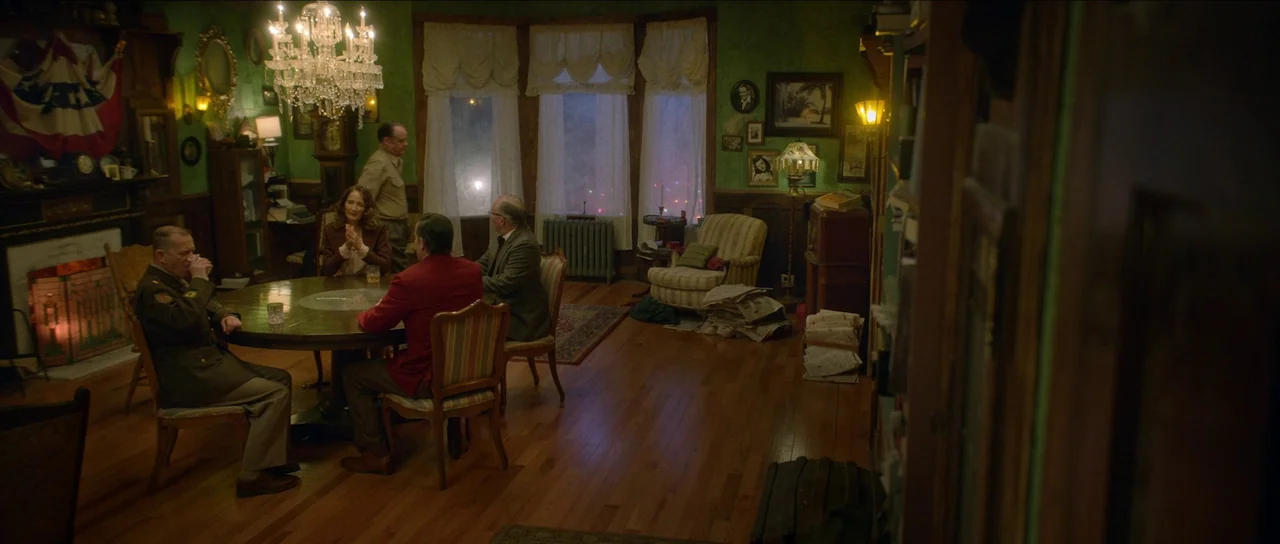Brooklyn 45 (2023) movie still: wide — A green-walled interior room features a crystal chandelier above a central dining table. Four adults…