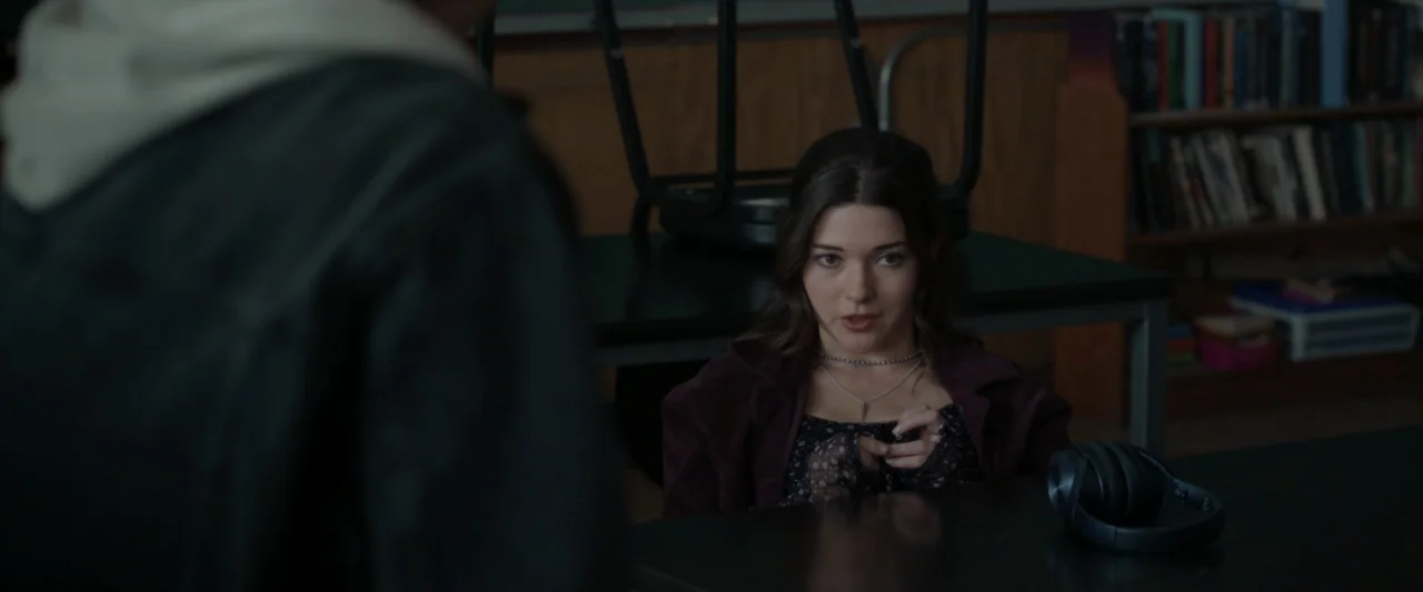 Clown in a Cornfield (2025) movie still: medium — A cluttered school room with desks and a bookshelf filled with textbooks. A young woman sits at a…