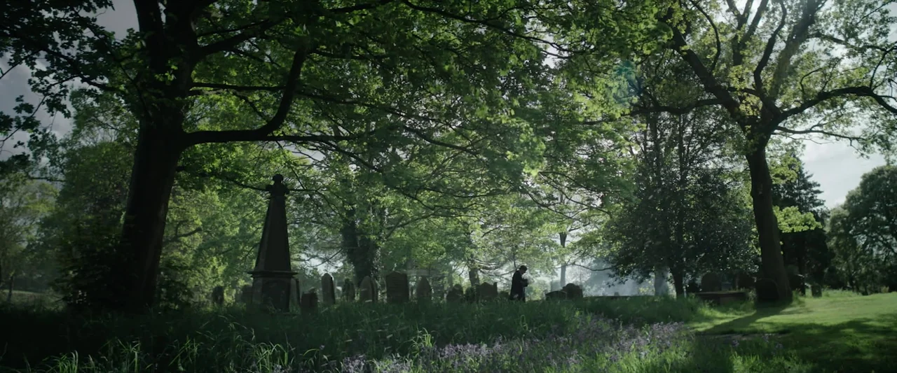 Nandor Fodor and the Talking Mongoose (2023) movie still: wide — A dense cemetery landscape features large mature trees, overgrown grass, and aged…