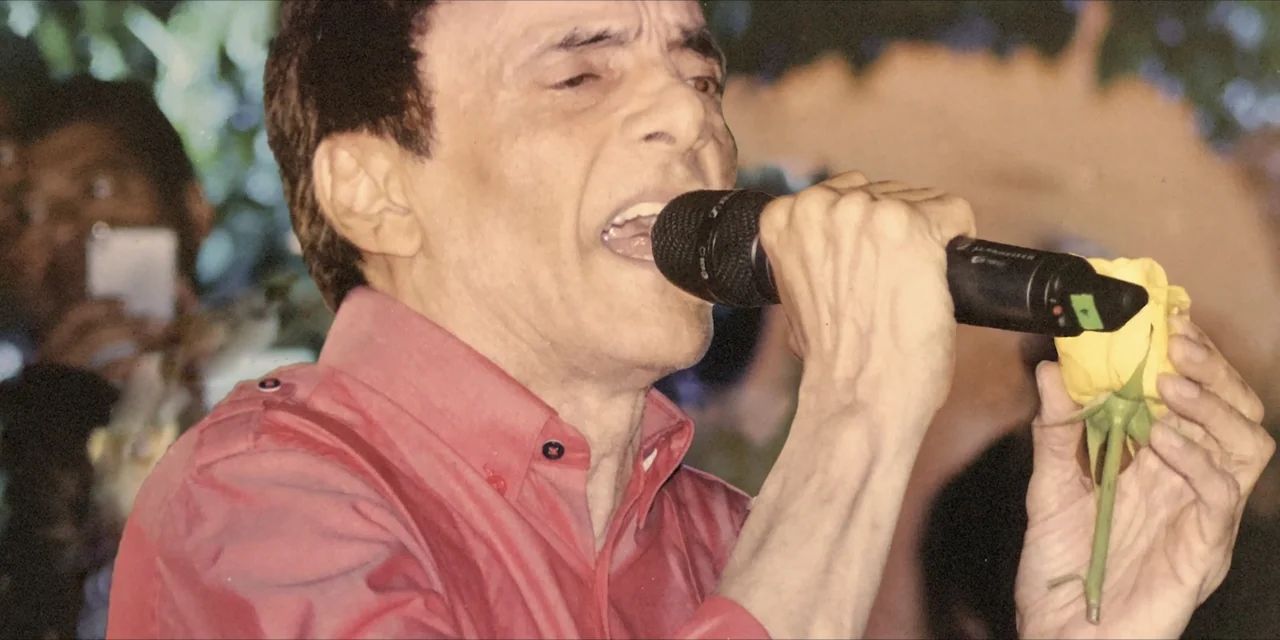 Leyenda Viva (2022) movie still: closeup — An outdoor performance area features a male singer under artificial ambient illumination. The performer…