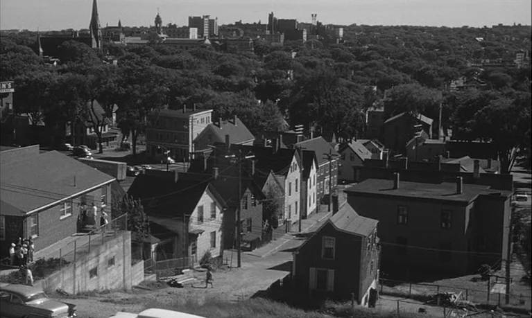 Lolita (1962) movie still: establishing — A high-angle view of a residential neighborhood with several small homes and trees. The urban skyline is…