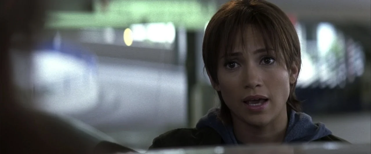 Enough (2002) movie still: closeup — A public indoor area features a blurred background with metallic and glass surfaces. A young woman with brown…