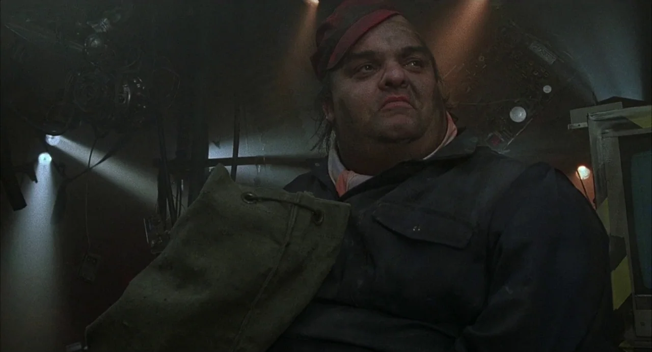Hardware (1990) movie still: medium — A dark, cramped interior space filled with wires and electronic equipment. A middle-aged man sits in the dim…