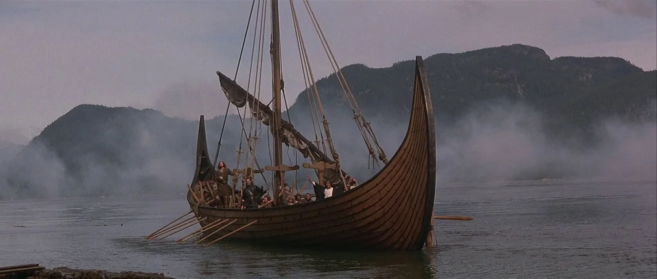 The 13th Warrior (1999) movie still: wide — A wooden Viking longship sits on calm water surrounded by misty mountains and hills. Several figures…