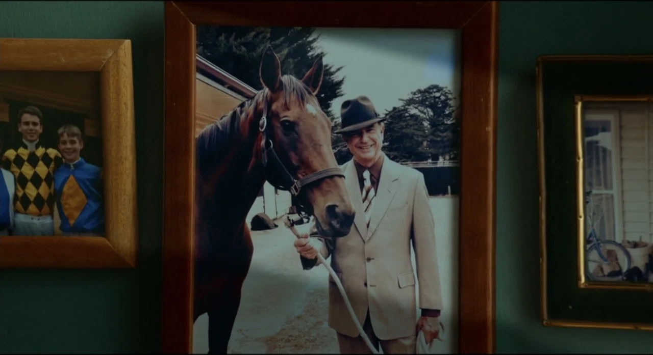Ride Like a Girl (2019) movie still: closeup — A close-up view of a framed photograph hanging on a dark teal wall. The photo depicts an older man…