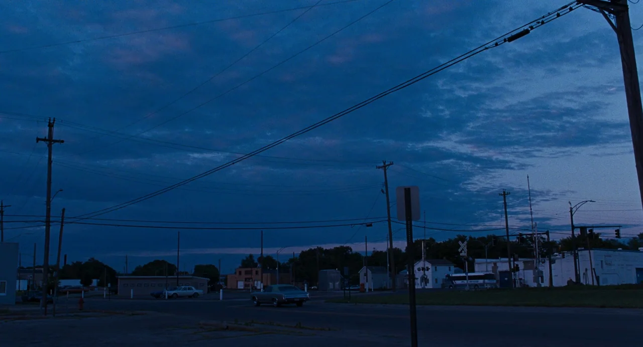 Bones and All (2022) movie still: establishing — An empty road intersection sits under a deep blue twilight sky with utility poles. A single vintage…