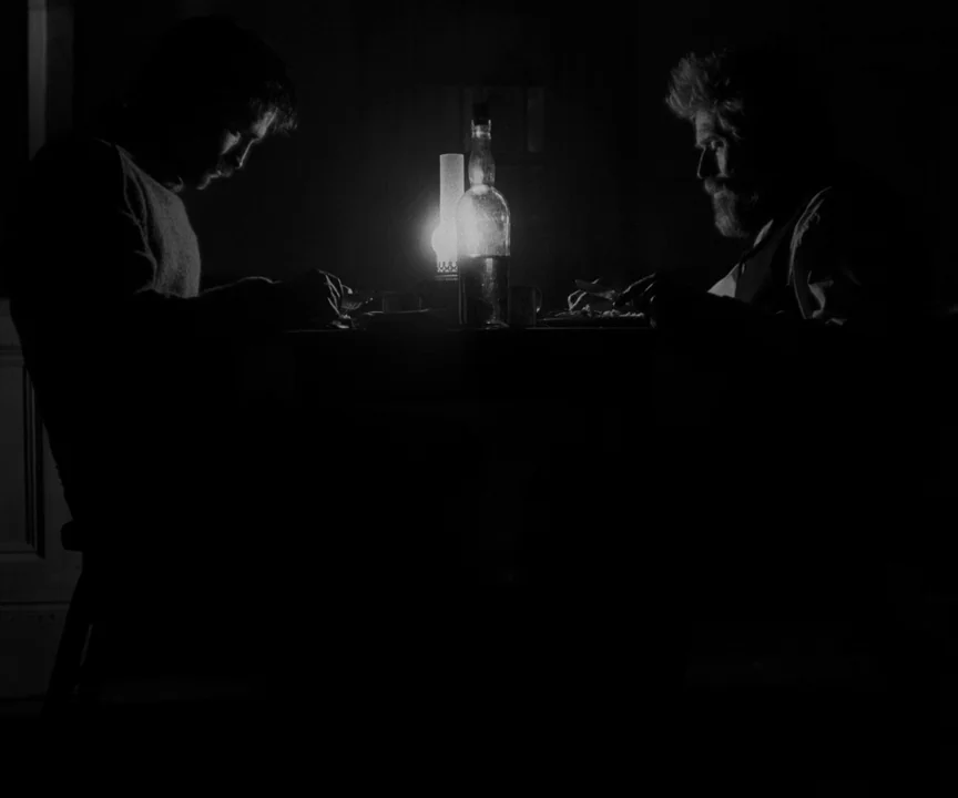 The Lighthouse (2019) movie still: medium — A dark, dimly lit room contains a table set for two individuals. The two men sit across from each other…