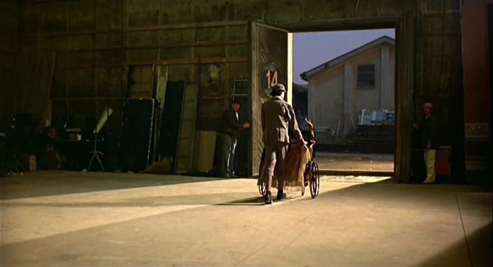 Intervista (1987) movie still: wide — An industrial building with large wooden doors open to an outdoor area with buildings. A man pushes a vintage…
