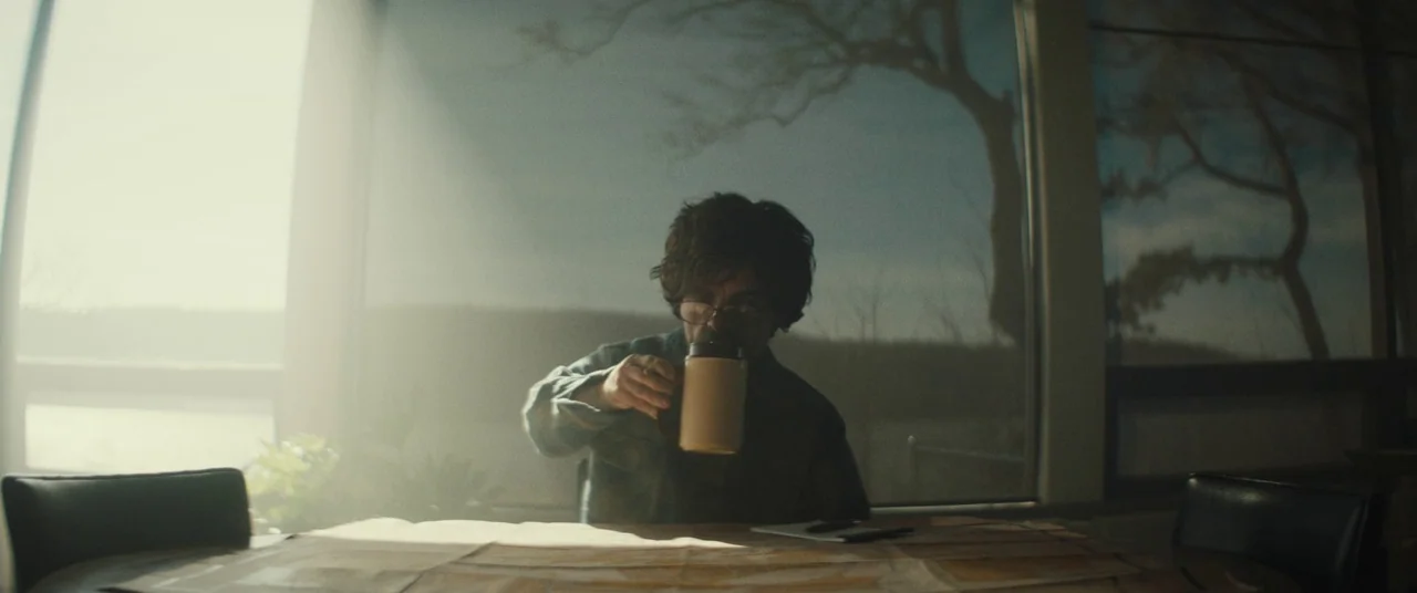 I Think We’re Alone Now (2018) movie still: medium — A man sits at a table inside a house with large windows facing outside. He lifts a tan travel…