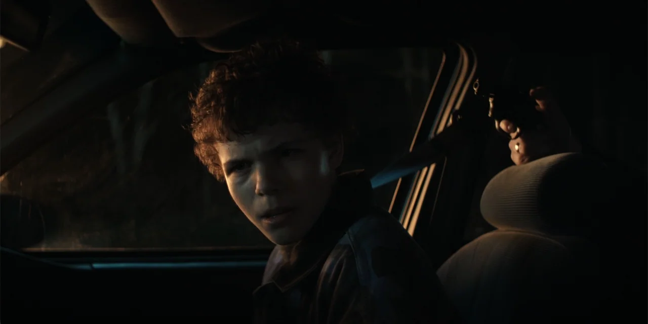 The Monkey (2025) movie still: medium — The interior of a dark vehicle at night with an obscured person behind. A teenage boy with curly hair turns…