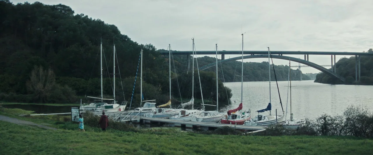 Vincent Must Die (2023) movie still: wide — A quiet marina features a floating wooden dock populated with several white sailboats. A large green…