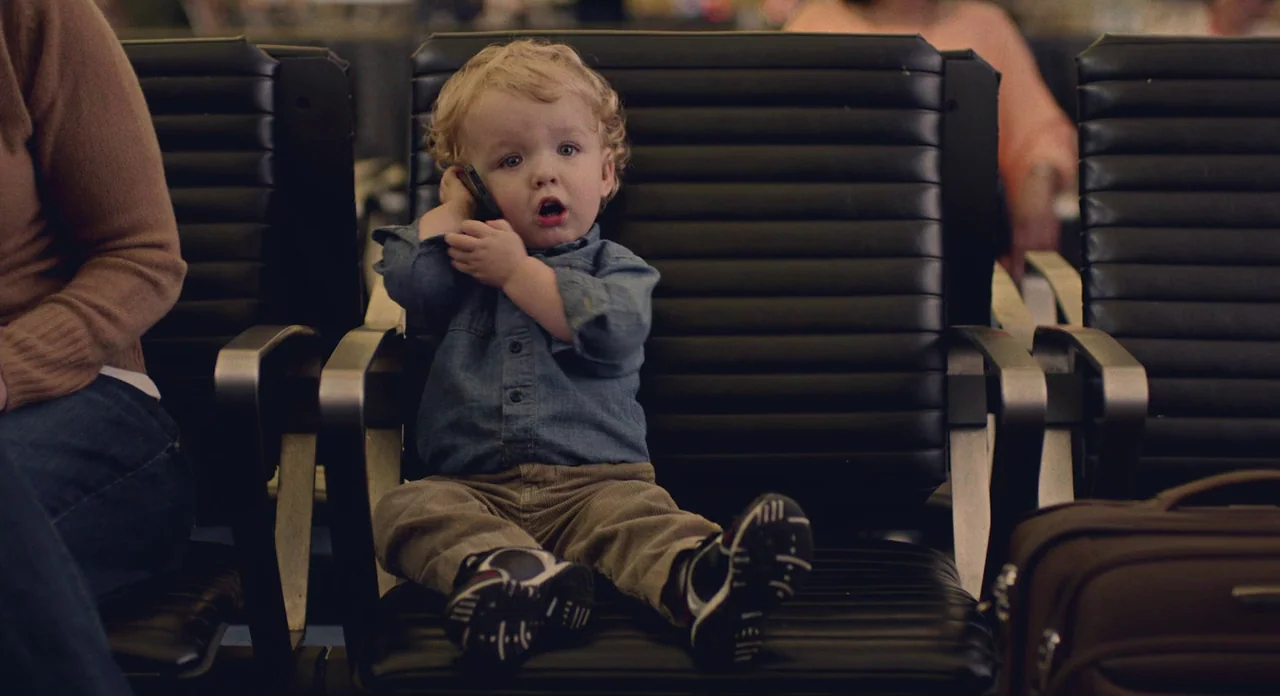 While We’re Young (2014) movie still: medium — A toddler sits on black padded seats in a large public transit waiting area. The child holds a black…