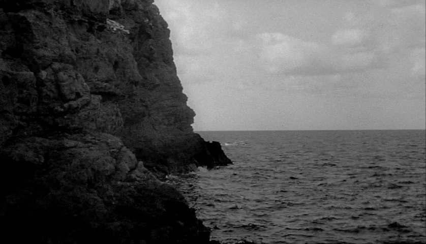L’Avventura (1960) movie still: establishing — A massive, jagged rock cliff face towers over dark, rippling ocean water. Low hanging clouds fill the…