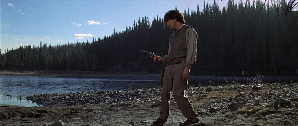 Little Big Man (1970) movie still: fullbody — A rocky riverbank is lined with a dense evergreen forest under a bright sky. A man stands on the shore…