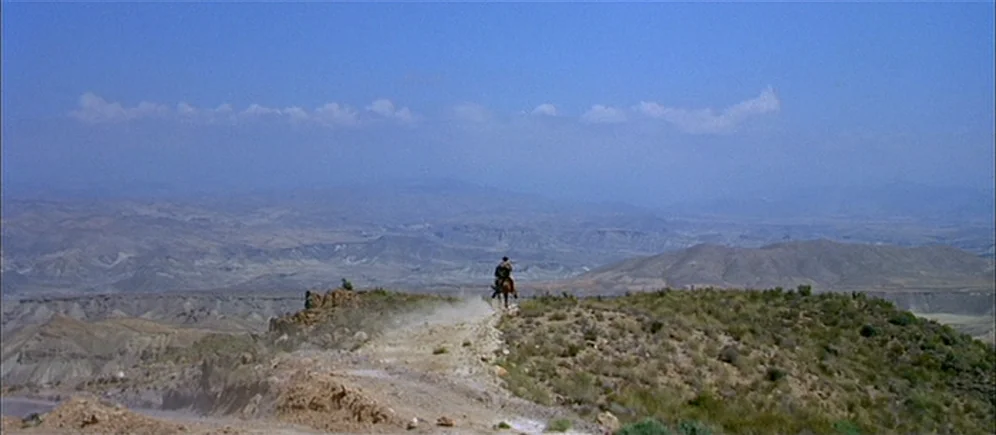 For A Few Dollars More (1965) movie still: wide — A vast arid desert landscape features rolling hills and distant mountains under a blue sky. A lone…