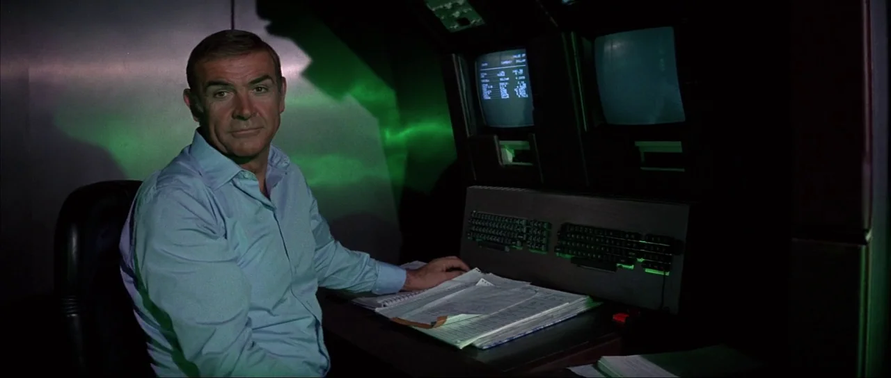 Never Say Never Again (1983) movie still: medium — A dark, modern technological workstation features glowing green computer screens and keyboards. A…