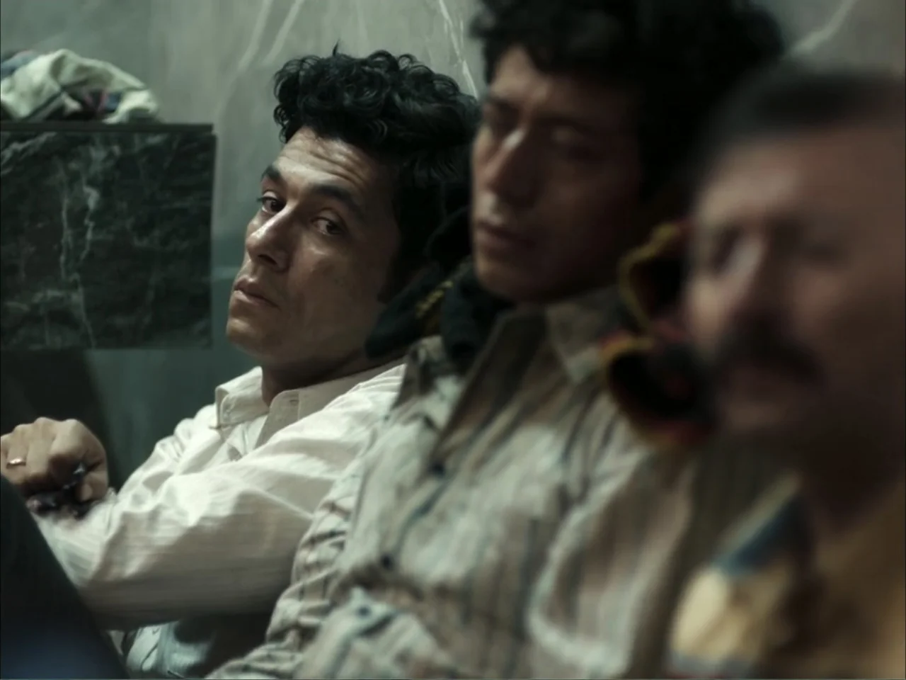 November (2025) movie still: medium — A dimly lit interior space with grey marble walls and three men sitting together. Three men sit side-by-side…