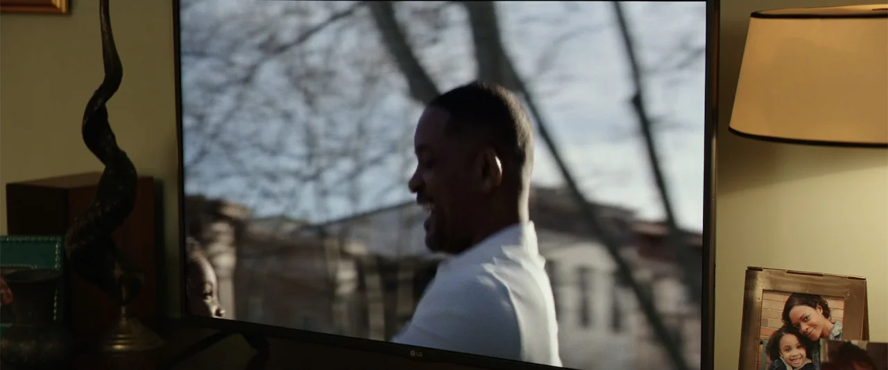 Collateral Beauty (2016) movie still: medium — A dimly lit room contains a television screen displaying an outdoor street scene. The screen shows a…