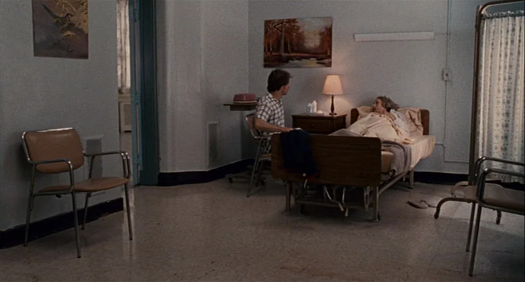Choke (2008) movie still: wide — A spare hospital room contains two chairs, a bed, and framed landscape artwork. A man in a plaid shirt sits beside…