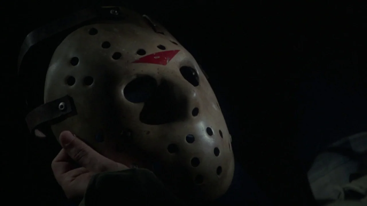 Friday the 13th Part VI: Jason Lives (1986) movie still: closeup — An off-white, worn hockey mask with a red triangle symbol is held against black.…