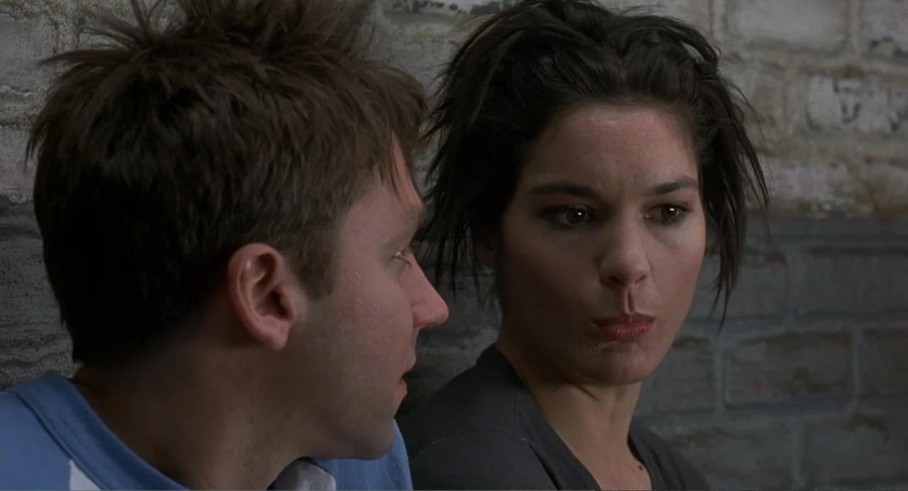 Wedding Daze (2006) movie still: medium — Two individuals sit against a painted brick wall in a dim outdoor urban area. The man and woman are…