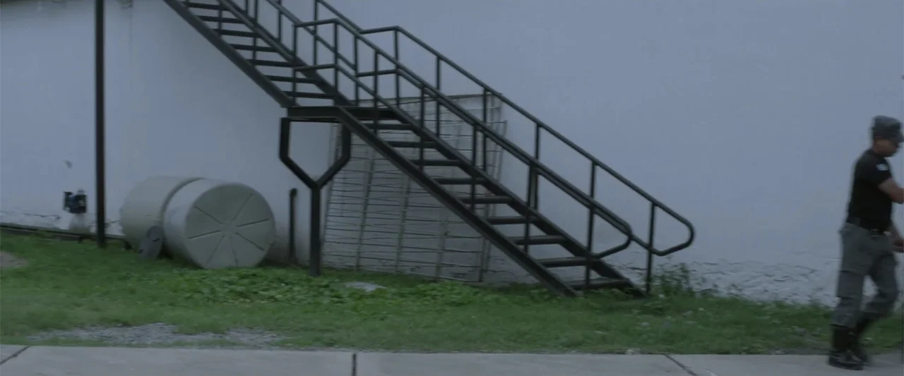 I Trust in You (2016) movie still: medium — A sterile alleyway features a white wall and a black metal exterior staircase. A man wearing a tactical…
