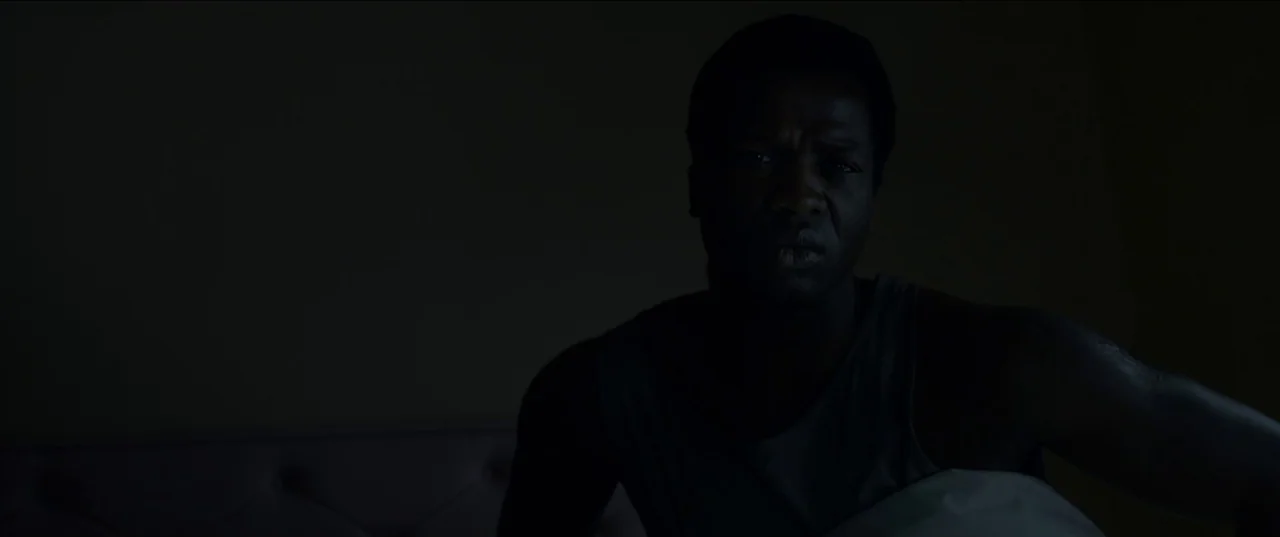 His House (2020) movie still: medium — A dark bedroom with an obscured headboard visible in the dim background. A man sits upright in bed staring…