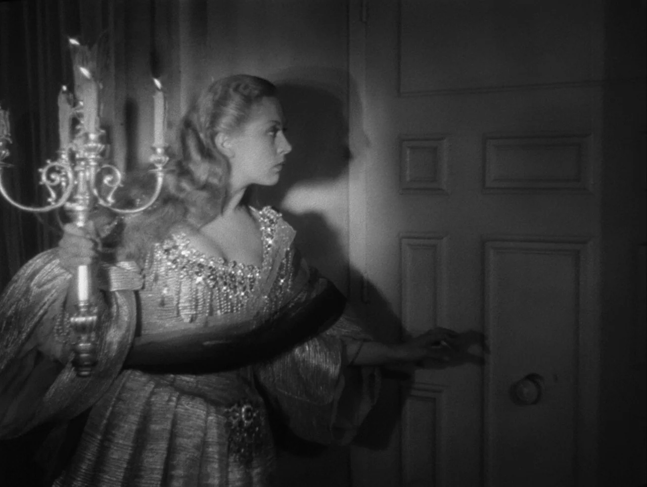 La Belle et la Bête (1946) movie still: medium — A woman stands in a dimly lit hallway next to a wooden door. She carries a silver candelabra and…