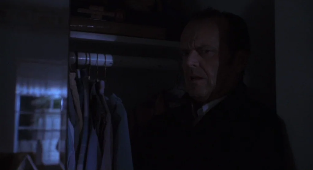 The Crossing Guard (1995) movie still: medium — A dark closet filled with hanging clothes occupies the foreground of a dim room. A man stares…