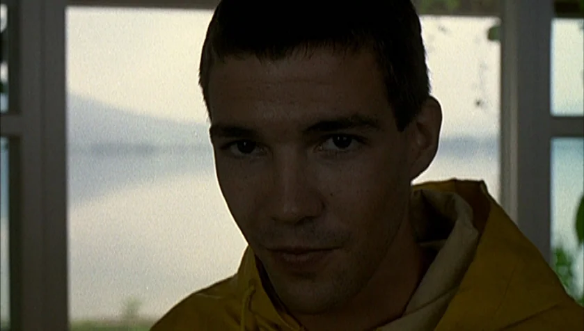 Funny Games (1997) movie still: closeup — An interior space with a large window looking out onto a body of water. A young man wearing a yellow…