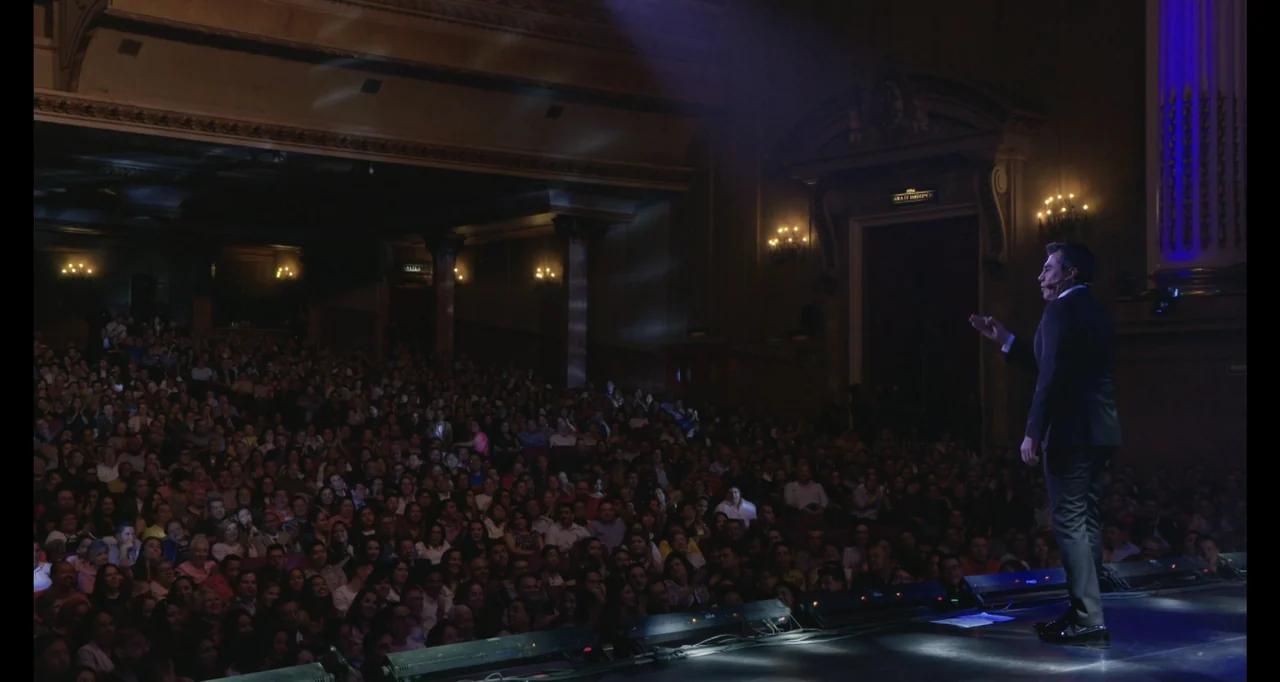 Paired Up (2019) movie still: wide — A large, dark theater auditorium is filled with a seated audience facing a stage. A man stands on the stage…
