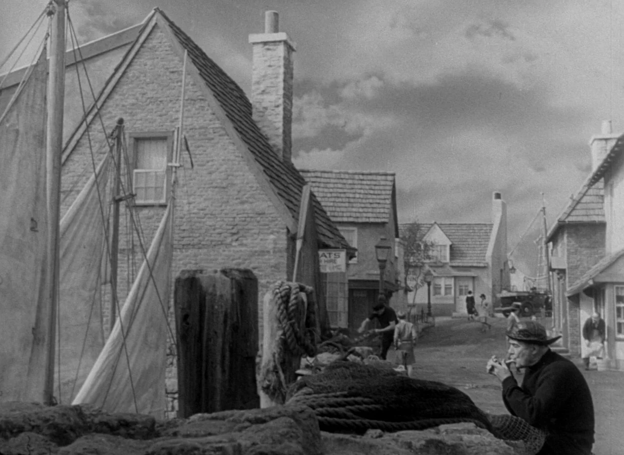 The Uninvited (1944) movie still: wide — A quiet coastal village street runs alongside stone buildings and a boat mooring area. A man sits in the…