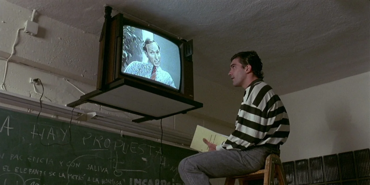 Law of Desire (1987) movie still: medium — A classroom features a green chalkboard with white writing and a mounted CRT television. A young man sits…