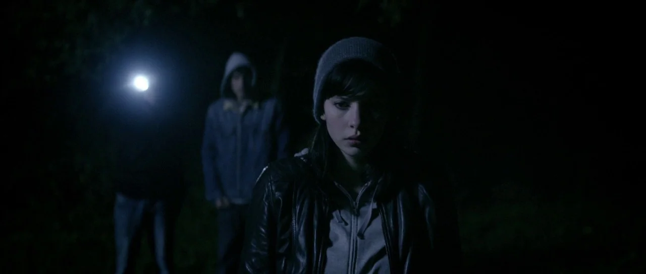 Livid (2011) movie still: medium — A dark outdoor space at night with dense vegetation obscured by shadows. A young woman in a beanie and leather…